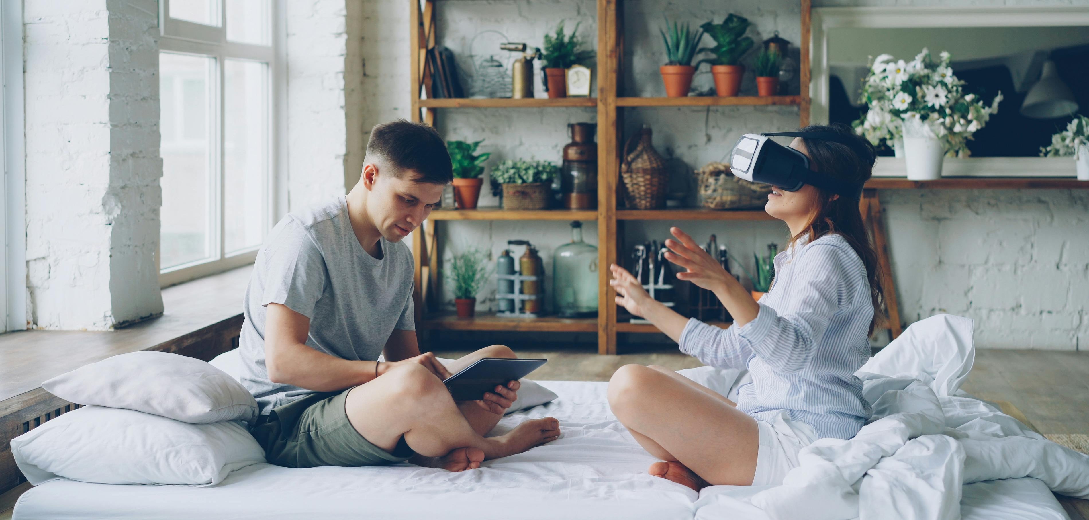 Man and woman sitting on a bed in a cozy room using smart devices